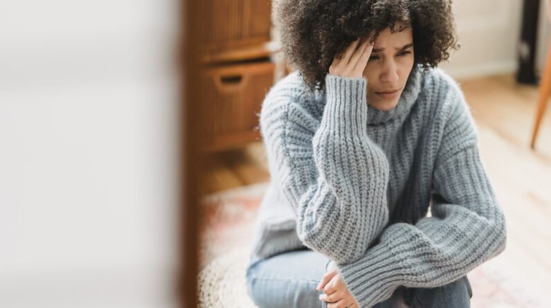 Young ethnic female in casual clothes sitting on pouf while crying with hand on head in light flat